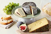 Various types of cheese on a wooden board and on a cake stand with figs, grapes and a cheese knife