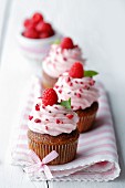 Raspberry cupcakes decorated with ribbon on a pink striped napkin with a bowl of raspberries in the background