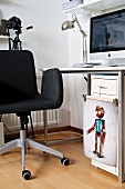 Dunkelgrauer Drehstuhl vor Schreibtisch mit Computer, Klammerbügel mit der bunten Illustration eines Roboters an Schubladenschrank
