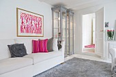 Femininer Wohnraum in Weiß und Grau mit pinkfarbenen Akzenten; beleuchtete Vitrinenschränke neben modernem Bild und Sofa