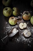 A halved apple and a flour scoop with flour sprinkled over the work surface and other apples in the background
