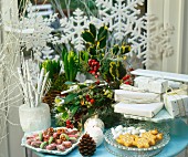 Weihnachtstisch mit Blumendeko, Geschenken, Kerzen und Weihnachtsplätzchen vor Fenster mit dekorativen Schneeflocken