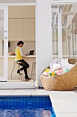 View from courtyard with pool and wicker armchair through open terrace door of woman in house interior