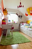 Schreibtisch und Stuhl in Weiß auf grünem Teppich gegenüber Bett mit Schubladen in Kinderzimmer unter dem Dach