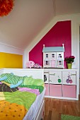 Bett mit bunt gemusterter Bettwäsche unter Dachschräge, im Hintergrund Regal mit Aufbewahrungsboxen und Puppenhaus vor pinkfarbener Wand