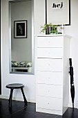 Narrow, white chest of drawers, black umbrella and stool on black tiled floor in hall