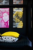 Detail of bed with yellow and black and white patterned scatter cushions on black throw and collection of posters on wall
