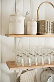 Storage jars, wicker basket and glasses on wooden shelves on white wooden wall