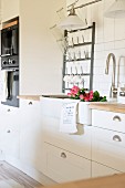 Detail of white, country-house kitchen with roses in sink and white metal bottle rack on wall