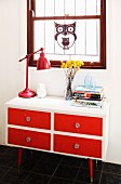 Red, retro table lamp on 50s-style, white chest of drawers with red drawer fronts below decorative ornament hung in window