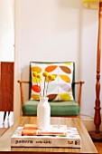 Simple vase of dried yellow flowers on stacked books and coffee table; 50s armchair with patterned scatter cushions in background