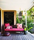 Bench with deep pink seat cushion and scatter cushions on wooden decking; tropical plants in background