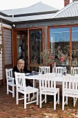 Frau auf Terrasse, weisslackierte Outdoorstühle und Tisch auf Ziegelboden vor Holzhaus mit raumhohen Terrassenfenstern