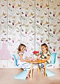 Kleine Mädchen auf hellblauen Stühlen am runden Tisch vor Einbauschrank mit Schmetterling-Tapete