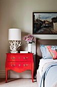 White table lamp on red vintage bureau next to bed below framed picture