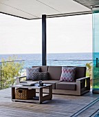 Relaxen auf überdachter Holzterrasse mit Aussicht auf den Pazifik, Coffee Table und passendes Sofa mit braunen Polstern und gemusterten Kissen vor Glasbrüstung