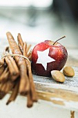 A Christmas apple decorated with a snow star next to cinnamon sticks and almonds on a table