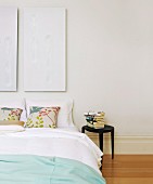 White diptych above double bed with floral scatter cushions next to stack of books on stool used as bedside table