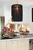 Pendant lamp with black, fabric lampshade above counter with sink and vase of flowers; illuminated gas stove in background