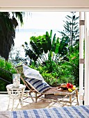 Sonnenliege aus Bambus und weisser Korbtisch auf sonniger Terrasse mit Blick auf Meer und tropische Pflanzen