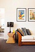 Framed retro posters above wicker sofa next to black table lamp on designer stool
