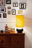 Table lamp with yellow fabric lampshade on wooden sideboard against white wainscoting and below framed photos