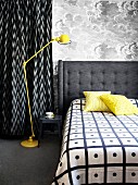 Doppelbett mit grau gepolstertem Kopfteil neben gelber Retro Stehleuchte vor Wand mit wolkiger Mustertapete