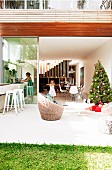 Christmas tree on sunny terrace in front of open, sliding glass door and view of family in interior