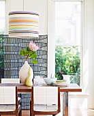 Protea flower in vase on dining table and chairs with white, upholstered backs below pendant lamp with striped lampshade
