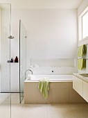 Helles Badezimmer mit Wandnische über der Wanne und Hängewaschtisch; seitlich eine mit Glaswänden abgetrennte, begehbare Dusche
