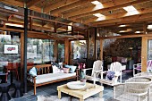 Terrace furnished with wooden bench, armchairs and wicker chairs in front of glass facade of house; wooden ceiling with skylights