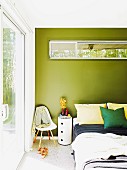 Rundes Schubladenmöbel und Klassikerstuhl als Kleiderablage neben Doppelbett; Oberlichtfenster mit Blick auf Bambushecke in grün getönter Wand