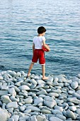 Kleiner Junge mit kurzer, roter Hose und Ball auf Kieselsteinen am Meeresufer