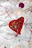 Red, hand-crafted love heart with colourful sequins and purple bauble on white, artificial Christmas tree