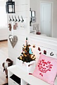 Pink cushion next to small, decorated Christmas tree in crate on white cloakroom bench in rustic hallway