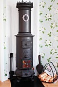Vintage, cylindrical wood-burning stove and basket of firewood in corner of room with romantic floral wallpaper
