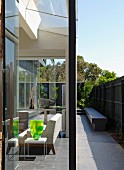 Glass walls create seamless transition between seating area in interior and narrow courtyard with stone bench against screen fence; decorative glass vase