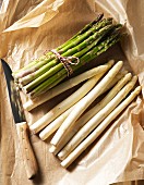 A bunch of green asparagus and loose white asparagus on a piece of paper with a knife