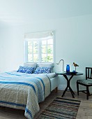 Französisches Bett mit gestreifter Tagesdecke und gemusterten Kissen vor Sprossenfenster, seitlich runder Beistelltisch mit Leuchte, minimalistisches, ländliches Ambiente