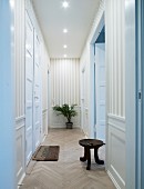 Schmaler Gang mit weissen, hohen Zimmertüren in traditionellem Stil an Wänden Streifentapete, rustikaler Holzschemel auf Fischgrätparkett
