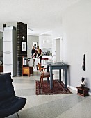 Vintage wooden chair at grey-painted table on Oriental rug in open-plan interior; woman in dining area in background