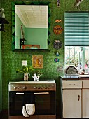 Moderner Küchenherd aus Edelstahl und Spiegel an Wand mit grünen Mosaikfliesen, seitlich Vintage-Spüle mit Unterschrank am Fenster