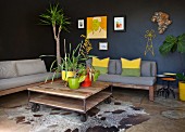 Plants in bright pots on rustic coffee table on castors in front of couches with seat cushions against black walls