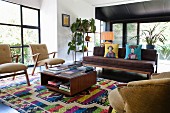 Retro-style armchair, couch and coffee table on brightly coloured rug in modern, open-plan interior