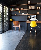 Classic yellow shell chair at minimalist desk below wooden shelves on black-painted wall; polished concrete and wooden floor