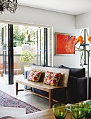 Simple wooden bench with ethnic cushions against backrest of black sofa in modern interior with view of garden through open balcony doors