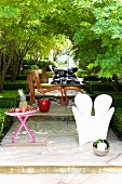 Various outdoor furniture on large, terraced steps in garden - mirrored ball on floor in front of white, plastic armchair, fruit bowl on table and rattan easy chair with matching footstool