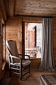 Blanket on rustic armchair next to French window in corner of wooden cabin