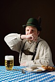 A stereotypical German man wearing lederhosen with beer and white sausage wiping his mouth