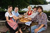 Five people sitting in a beer garden at a table with tankards and pretzels